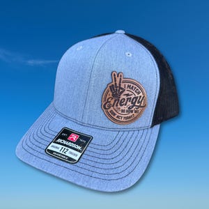 May include: A light blue Richardson 112 trucker hat with a black mesh back. The hat features a brown leather patch with the words "I Match Energy, So How We Gon' Act Today?" and a peace sign. Adjustable fit.