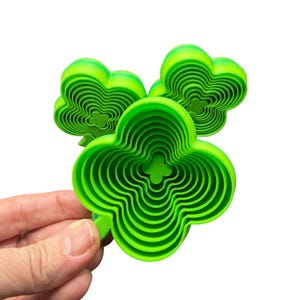 May include: Three bright green, layered clover-shaped objects. Each clover has multiple concentric layers, creating a 3D effect. The objects are held against a white background, highlighting their vibrant color and unique design. The clover shapes are a symbol of good luck.