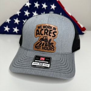 May include: A gray and black trucker hat with a brown leather patch that says "We work in acres not hours". The patch features a black silhouette of a tractor and wheat stalks. The hat also has a black Richardson 112 label.