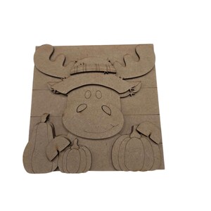 May include: A light brown wooden cutout sign featuring a moose wearing a hat with a plaid pattern. The moose is positioned above three pumpkins of varying shapes and sizes. The entire design is layered on a rectangular wooden board background.