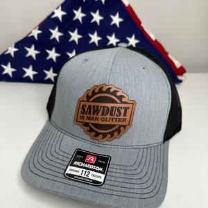 May include: A gray and black trucker hat with a brown leather patch that says "Sawdust is Man Glitter". The hat is adjustable and has a Richardson 112 logo.