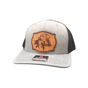 May include: Gray trucker hat with a black mesh back and a brown leather patch. The patch features a Labrador Retriever with a bird in its mouth. The hat has a curved brim and the text "RICHARDSON 112" on the label.
