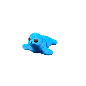 May include: A small, light blue seal figurine with black eyes and a smiling expression. The seal is lying down, with its flippers spread out. The figurine is made of a smooth material and is set against a white background.