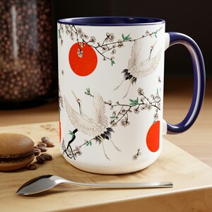 May include: White ceramic mug with a blue rim and handle. The mug features a repeating pattern of red circles, white cranes, and black branches with white blossoms.