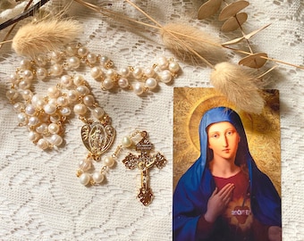 Puede incluir: Un rosario de perlas con detalles dorados y un crucifijo dorado. También hay una tarjeta religiosa con la imagen de una mujer con un velo azul. Los elementos están dispuestos sobre una superficie de encaje blanco.