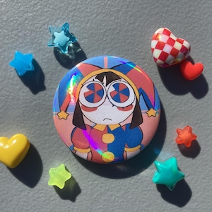 May include: A round button with a cartoon jester design, surrounded by small star and heart-shaped embellishments. The button features a colorful, holographic effect. The embellishments are in various colors, including blue, yellow, red, and orange.