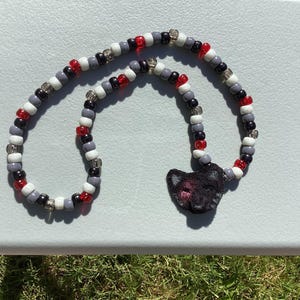 May include: A beaded necklace with a black cat pendant. The necklace features a pattern of red, white, gray, and black beads. The cat pendant is black with pink embroidered details. The necklace is lying on a light gray surface.