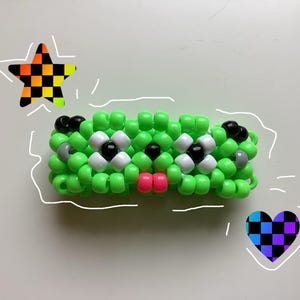 May include: A green beaded bracelet shaped like a cartoon face with black, white, and gray beads for eyes, and a pink bead for the mouth. A star and heart with checkered patterns are drawn on the background.