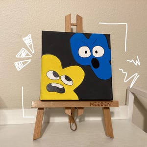 May include: A small canvas painting featuring two cartoon characters against a black background. One character is yellow, the other is blue. The painting is displayed on a miniature wooden easel. The word "MEEDEN" is visible on the easel.