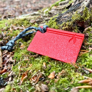 Może przedstawiać: Czerwony plastikowy klin do campingu z fakturowaną powierzchnią i przymocowanym czarno-szarym sznurkiem. Klin leży na podkładce z zielonego mchu.