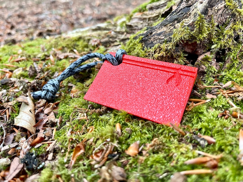 Może przedstawiać: Czerwony plastikowy klin do campingu z fakturowaną powierzchnią i przymocowanym czarno-szarym sznurkiem. Klin leży na podkładce z zielonego mchu.