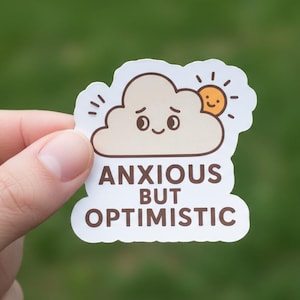May include: A sticker featuring a cartoon cloud with a sad face and a smiling sun. The text "ANXIOUS BUT OPTIMISTIC" is printed below the cloud. The sticker is held against a blurred green background.