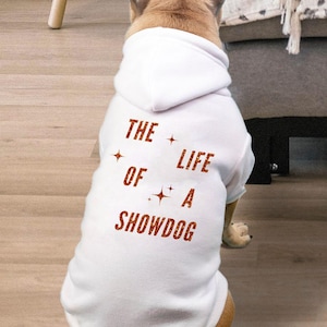 Peut inclure: Un sweat à capuche blanc pour chien avec le texte "THE LIFE OF A SHOWDOG" en rouge. Le chien est brun et beige, assis sur un sol en bois. Le sweat à capuche a une capuche.