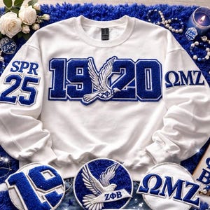 Peut inclure: Sweat-shirt blanc avec broderies bleues. Il affiche "1920" avec un emblème de colombe à l'avant, et "SPR 25" et "ΩMZ" sur les manches. Des écussons supplémentaires avec des motifs similaires sont également visibles.