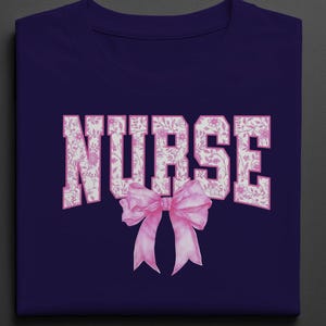 以下が含まれることがあります： ネイビーブルーのTシャツ。白いブロック体の「NURSE」の文字はピンクの花柄で埋められています。ピンクの水彩画のリボンが単語の下に配置されています。