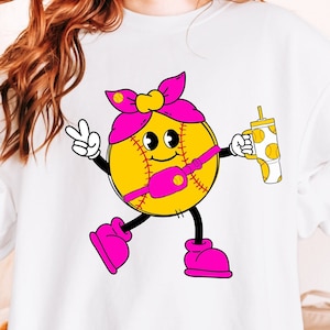 Può includere: Felpa bianca con un personaggio di softball dei cartoni animati. La palla da softball ha un fiocco rosa, una cintura e scarpe, e tiene in mano una bevanda e fa il segno della pace. Il personaggio ha un volto sorridente e arti neri.