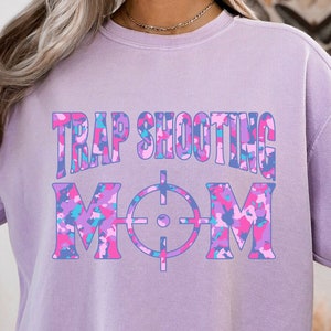 May include: A lavender sweatshirt with a camouflage print design. The text "TRAP SHOOTING MOM" is printed on the front of the sweatshirt.