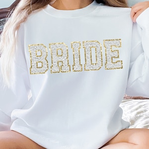 Puede incluir: Sudadera blanca con la palabra "BRIDE" en letras doradas con purpurina.