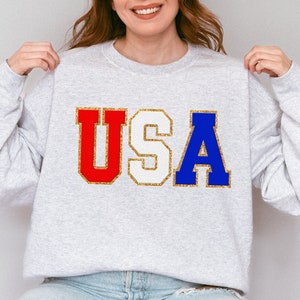 以下が含まれることがあります： 前面に「USA」という文字が赤、白、青の文字で金色に輝くアクセントでプリントされたグレーのスウェットシャツです。