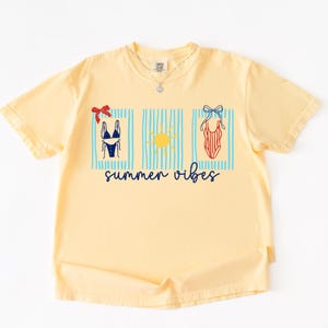 Op de afbeelding: Een lichtgeel t-shirt met een grafisch ontwerp met een blauw-witte bikini, een gele zon en een rood-wit badpak. De woorden "summer vibes" staan in blauwe letters eronder.