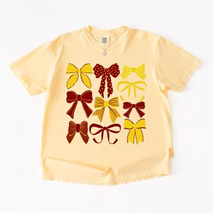 Puede incluir: Camiseta amarilla clara con un diseño de lazos en amarillo y burdeos. Los lazos tienen diferentes estilos, algunos con lunares. La camiseta tiene mangas cortas y cuello redondo. El diseño está centrado.