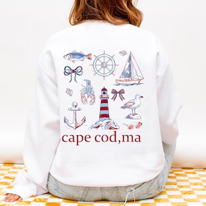 Puede incluir: Sudadera blanca con ilustraciones náuticas en rojo y azul, incluyendo un faro, un velero y un ancla. El texto "cape cod, ma" está impreso debajo. Una prenda cómoda e informal.