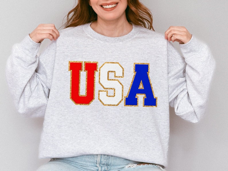 May include: A gray sweatshirt with the word "USA" printed on the front in red, white, and blue letters with gold glitter accents.