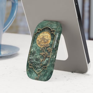 May include: A green and gold phone stand with a decorative design of a tree and moon. The stand is made of a textured material and has a raised edge to hold a phone in place.