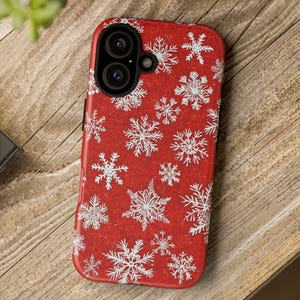 May include: A red phone case with a white snowflake pattern. The case has a dual-lens camera cutout and is resting on a wooden surface. The design features various sizes of snowflakes.