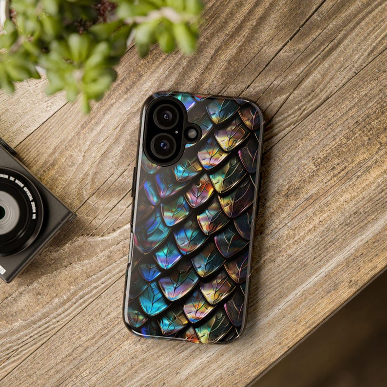 May include: A smartphone case featuring a repeating pattern of iridescent, metallic scales in shades of deep blue, purple, and gold. The design covers the entire back of the case, which includes a cutout for a dual-lens camera system.