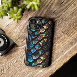 May include: A smartphone case featuring a repeating pattern of iridescent, metallic scales in shades of deep blue, purple, and gold. The case has a cutout for a dual-lens camera system on the upper left corner and a black border surrounding the edges.