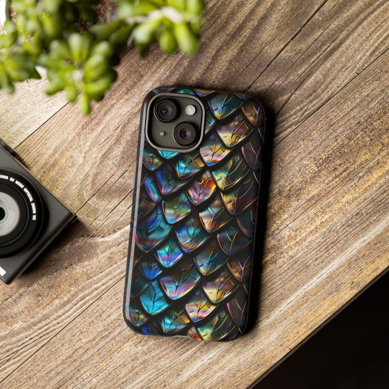 May include: A smartphone case featuring a repeating pattern of iridescent, metallic scales in shades of deep blue, purple, and gold. The case has a cutout for a dual-lens camera system on the upper left corner and a black border surrounding the edges.