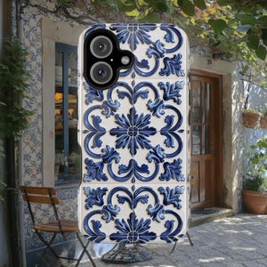 Puede incluir: Una funda para teléfono blanca y azul con un patrón geométrico inspirado en los azulejos portugueses tradicionales. La funda tiene una protuberancia de cámara elevada y un recorte para el puerto de carga del teléfono.