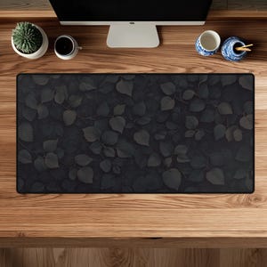 May include: A rectangular desk mat featuring a dark, monochromatic pattern of overlapping leaves in shades of charcoal and black. The mat has a solid black border and is positioned on a wooden surface below a computer monitor and desk accessories.