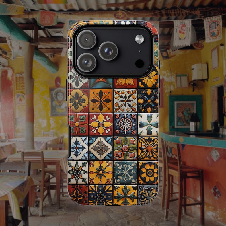 Op de afbeelding: Een smartphonehoesje met een raster van kleurrijke, vierkante tegels met ingewikkelde bloem- en geometrische patronen in tinten geel, oranje, blauw, groen en wit. Het hoesje heeft een zwarte camera-uitsparing aan de bovenkant met drie lenzen.