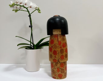 Może przedstawiać: Drewniana lalka kokeshi z czarną peruką i czerwono-pomarańczową kimonową sukienką. Lalka stoi na białym tle przed białą orchideą.