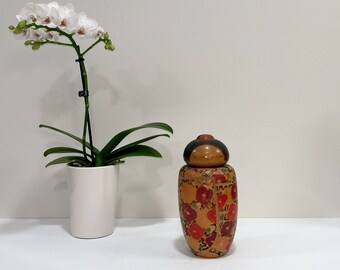 Może przedstawiać: Biała orchidea w białej ceramicznej doniczce obok drewnianego pojemnika w kształcie lalki z czerwonymi i czarnymi wzorami kwiatowymi.