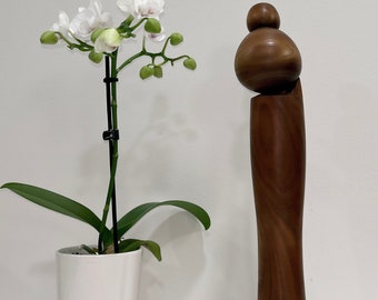 May include: A wooden sculpture of a person with a rounded head and a long, slender body. The sculpture is brown and has a natural wood finish. A white orchid plant is in a white ceramic pot to the left of the sculpture.