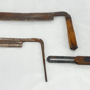 May include: Three antique woodworking tools are displayed. A large, curved drawknife with wooden handles, a smaller drawknife, and a metal and wood angle gauge. The tools have a rustic, aged appearance.