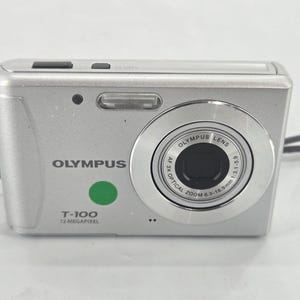 May include: Silver Olympus T-100 digital camera with a black lens and a green button. The camera has a 12-megapixel resolution and a zoom lens. A black strap is attached to the side.
