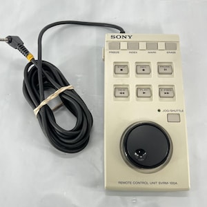 May include: A beige Sony Remote Control Unit SVRM-100A with black buttons and a jog/shuttle dial. It has a black cable and a 3.5mm jack. The text on the unit includes "FREEZE", "INDEX", "MARK", "ERASE", "STOP", "REC", "PAUSE", "REW", "PLAY", and "FFWD".