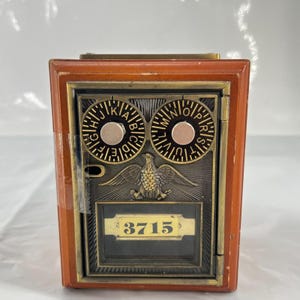 May include: Vintage postal box with a wooden frame and metal front. The metal front features two circular dials with letters, an eagle emblem, and the number "3715" in a rectangular frame. The box has a brown and gold color scheme.
