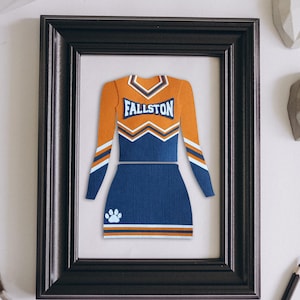 May include: A framed paper cutout of a cheerleading uniform featuring an orange long-sleeve top with a blue V-neck and the text FALLSTON, paired with a blue skirt. The design includes white and orange stripes and a white paw print on the skirt.