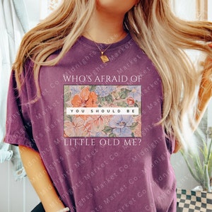 Peut inclure: Un t-shirt violet avec le texte "WHO'S AFRAID OF YOU SHOULD BE LITTLE OLD ME?" sur un motif floral. Le motif floral présente des fleurs orange, bleues et roses. Le t-shirt est de couleur unie et a une coupe décontractée.