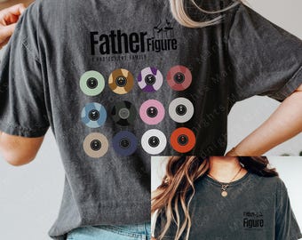 May include: Dark gray t-shirt with the text "Father Figure" and the phrase "I PROTECT THE FAMILY". The back of the shirt features a design of twelve colorful vinyl records. The shirt is a casual, unisex style.