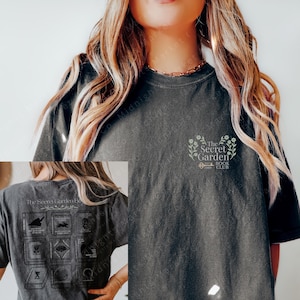 May include: A black t-shirt with a graphic design on the front that says "The Secret Garden Book Club". The back of the shirt has a graphic design of a book with a key and the words "The Secret Garden Book Club".