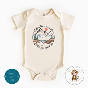 May include: A cream coloured baby onesie with a graphic of a mountain range with a lake and a red sun. The text "Adventure awaits let's go find it" is written in a circle around the graphic.