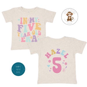 Peut inclure: T-shirt beige clair pour enfant avec l'inscription "IN MY FIVE YEAR OLD ERA" dans une police rétro colorée. Le dos du t-shirt affiche "HAZEL 5" avec des fleurs, des cœurs et un signe de paix.