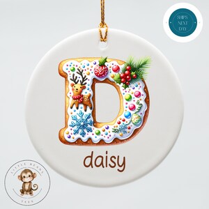 May include: A white ceramic Christmas ornament with a gingerbread letter D decorated with colorful sprinkles, a reindeer, holly, and a snowflake. The ornament has the name "daisy" written below the letter.