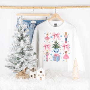 Op de afbeelding: Witte kersttrui met een feestelijk ontwerp met een ballerina, een notenkraker, een kerstboom en een cadeaudoos. Het ontwerp is in roze, blauw en goud.
